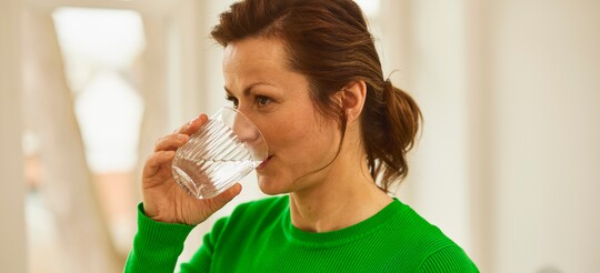 Frau trinkt Wasser als Hausmittel gegen Halsschmerzen – Hydratation zur Linderung. Frau in grünem Pullover trinkt ein Glas Wasser in einem hellen Raum. Das Trinken von Wasser ist ein bewährtes Hausmittel zur Linderung von Halsschmerzen und zur Förderung der Feuchtigkeit im Hals.