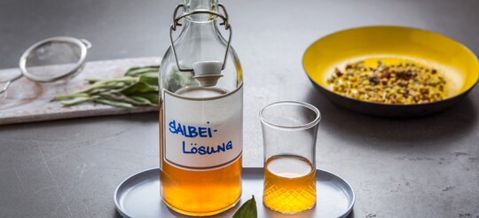 Glasflasche mit Salbei-Lösung und passendem Glas auf einem Tablett, daneben frische Salbeiblätter und eine Schale mit getrockneten Kräutern. Hausmittel zur Linderung von Halsbeschwerden.