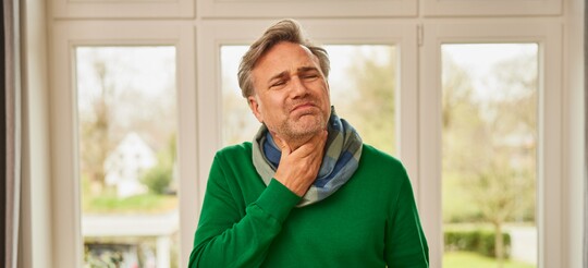 Linderung bei Halsschmerzen – GeloRevoice® bietet schnelle Hilfe. Ein Mann in einem grünen Pullover fasst sich mit schmerzverzerrtem Gesicht an den Hals, als Zeichen von Halsschmerzen, während er in einem hellen Raum mit großen Fenstern steht.