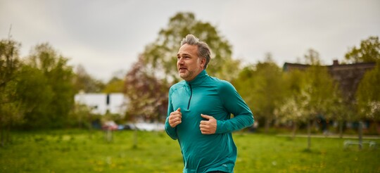 Mann beim Joggen im Park – Sport und Bewegung bei Halsschmerzen. Ein Mann mittleren Alters joggt im Freien auf einem Pfad durch eine grüne Parklandschaft. Er trägt ein türkisfarbenes Langarmshirt und wirkt entspannt. Die Szene zeigt die Verbindung von sportlicher Aktivität und der Vorsorge bzw. Linderung von Halsschmerzen durch Bewegung.