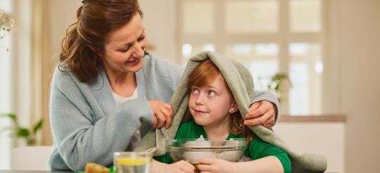 Eine Mutter hilft ihrem Kind beim Inhalieren über einer Schüssel mit heißem Wasser. Das Kind trägt ein grünes Oberteil und hat ein Handtuch über den Kopf gelegt. Auf dem Tisch steht eine Tasse heißes Wasser mit Zitronenscheiben. Die Szene zeigt ein traditionelles Hausmittel gegen Heiserkeit und Erkältungssymptome.