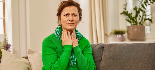 Eine Frau in einem grünen Pullover und Schal sitzt auf einem Sofa und hält sich den schmerzenden Hals. Sie wirkt besorgt, was auf Halsschmerzen oder Heiserkeit hinweist. Im Hintergrund stehen Pflanzen und Kissen, was eine häusliche und entspannte Atmosphäre schafft.