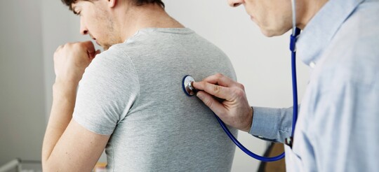 Ärztliche Untersuchung bei Husten – Unterstützung der Stimme mit GeloRevoice® Arzt untersucht mit einem Stethoskop einen hustenden Mann auf Atemprobleme, möglicherweise wegen einer Erkältung oder Bronchitis.