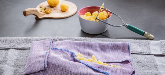 Gekochte und zerdrückte Kartoffeln auf einem Küchenhandtuch vorbereitet, mit Kartoffelstampfer und Schale auf einem Schneidebrett im Hintergrund, als Vorbereitung für ein Kartoffelwickel-Hausmittel gegen Halsbeschwerden.