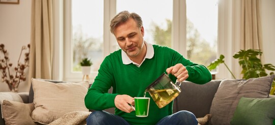 Mann gießt sich Kräutertee ein – Hausmittel gegen Reizhusten. Ein Mann mittleren Alters sitzt auf einem Sofa und gießt sich eine Tasse Kräutertee aus einer Teekanne ein. Er trägt einen grünen Pullover, und im Hintergrund sind Kissen und Pflanzen zu sehen. Die Szene vermittelt die Anwendung von Hausmitteln wie Kräutertee zur Linderung von Reizhusten.