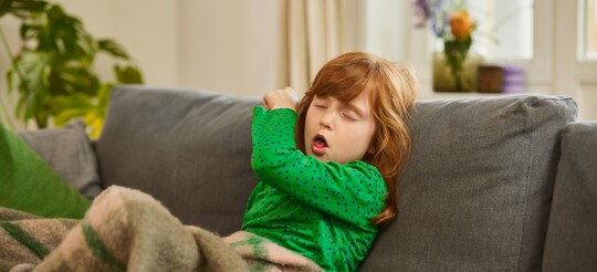 Hustendes Mädchen auf Sofa – GeloRevoice® zur Linderung von Reizhusten bei Kindern. Ein rothaariges Mädchen hustet in ihren Ellenbogen, während es auf einem Sofa sitzt, in eine Decke gehüllt.