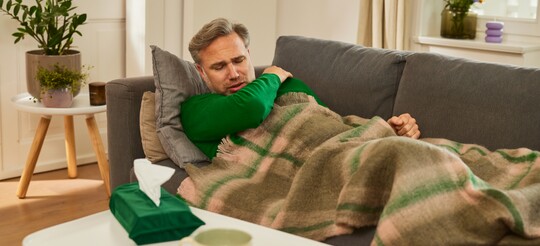 Kranker Mann mit Halsschmerzen, eingehüllt auf dem Sofa – Linderung mit Hausmitteln. Ein Mann liegt krank auf einem Sofa, zugedeckt mit einer Decke, und hält sich den schmerzenden Hals. Auf dem Tisch vor ihm stehen eine Tasse Tee, Zitronenscheiben und eine Packung Taschentücher.