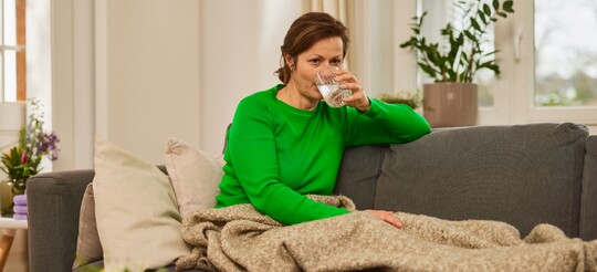 Frau trinkt Wasser auf dem Sofa – Flüssigkeitszufuhr als Teil der Stimmhygiene. Frau in grünem Pullover sitzt auf einem Sofa und trinkt ein Glas Wasser. Sie ist in eine beige Decke gehüllt, und im Hintergrund stehen Pflanzen, was eine ruhige und entspannte Atmosphäre schafft. Die Szene deutet auf Stimmhygiene und die Wichtigkeit der Flüssigkeitszufuhr für eine gesunde Stimme hin.