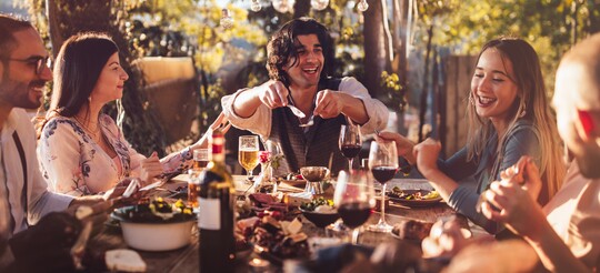 Eine Gruppe von Freunden sitzt bei Sonnenuntergang an einem festlich gedeckten Tisch im Freien und genießt ein gemeinsames Abendessen. Sie lachen, unterhalten sich und teilen Speisen in entspannter, fröhlicher Atmosphäre unter Lichterketten.