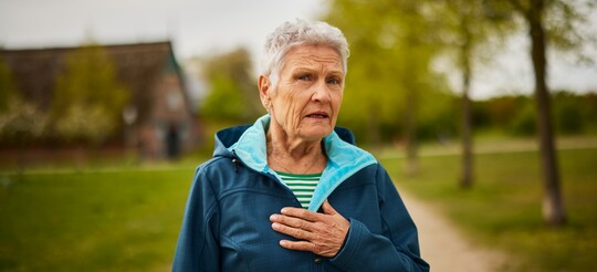 Ältere Frau mit Anzeichen von Presbyphonie – Stimmverlust und Stimmstörungen im Alter erkennen und mit GeloRevoice® unterstützen Ältere Frau im Freien, die besorgt ihre Hand auf die Brust legt, möglicherweise als Ausdruck stimmlicher Beschwerden im Alter. Die Szene thematisiert die Herausforderungen durch Presbyphonie, bei der die Stimme brüchig und weniger kräftig wird, oft begleitet von einem typischen Zittern des Kehlkopfes.