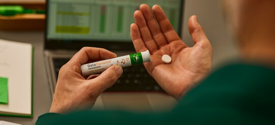 GeloRevoice® Halstabletten: Für langanhaltende Linderung bei Heiserkeit und Halskratzen im Arbeitsalltag Person hält eine GeloRevoice®-Packung und eine Lutschtablette in der Hand, vor einem Laptop auf dem Schreibtisch, bereit zur Einnahme für eine beruhigte Stimme.
