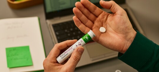 GeloRevoice® Halstablette zur Linderung von Halsbeschwerden Hand hält eine GeloRevoice® Halstablette und eine Tablettendose über einem geöffneten Laptop.