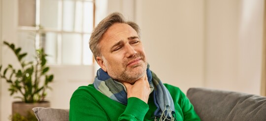 Schmerzen im Hals und Heiserkeit – Ursachen und Linderung bei Aphonie Mann mittleren Alters in grünem Pullover sitzt auf einem Sofa, hält sich den schmerzenden Hals und wirkt gequält. Er trägt einen Schal und scheint unter Heiserkeit oder Stimmverlust zu leiden.