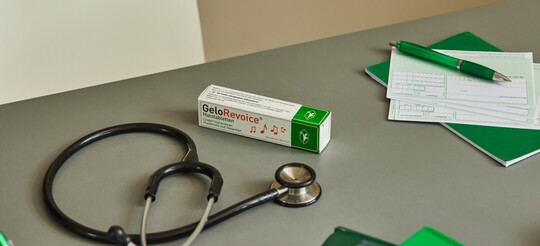Auf einem Schreibtisch in einer Arztpraxis liegen eine Packung GeloRevoice Halstabletten, ein Stethoskop, grüne Notizblöcke und ein Rezeptblock mit einem grünen Stift. Die Szene vermittelt eine professionelle medizinische Umgebung zur Behandlung von Heiserkeit.