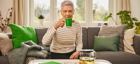 Ältere Frau entspannt mit einer Tasse Kräutertee zur Beruhigung von Hals und Stimme. Ältere Frau sitzt auf einem grauen Sofa in einem gemütlichen Wohnzimmer und trinkt aus einer grünen Tasse, auf dem Tisch vor ihr steht eine Glaskanne mit Kräutertee.