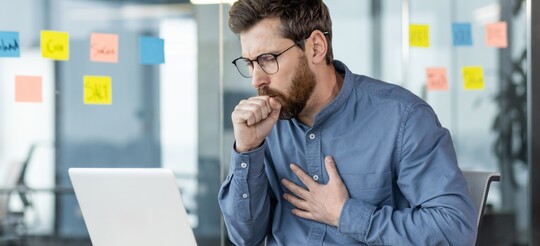 Stimmprobleme und Husten: Belastung der Stimme durch alltäglichen Stress – Tipps auf Gelorevoice.de. Ein Mann mit Brille und blauem Hemd sitzt an einem Schreibtisch mit einem Laptop und hält sich die Brust, während er hustet. Im Hintergrund ist ein modernes Büro mit Glaswand und bunten Haftnotizen zu sehen.