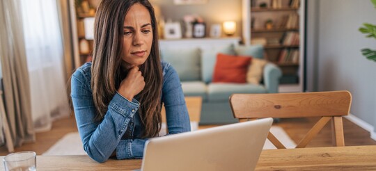 Stimmstörungen im Alltag und Beruf – Herausforderungen bei Dysphonie bewältigen. Frau in Jeansjacke sitzt nachdenklich an einem Holztisch und blickt auf den Bildschirm eines Laptops. Die Szene zeigt eine ruhige Umgebung mit einem gemütlichen Wohnzimmer im Hintergrund. Symbolische Darstellung der Herausforderung, Stimmstörungen wie Dysphonie im beruflichen oder alltäglichen Leben zu bewältigen, insbesondere für Personen, die ihre Stimme intensiv nutzen.
