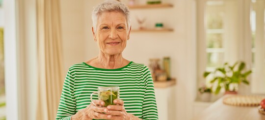 Ältere Frau trinkt Kräutertee – Hausmittel zur Linderung von Halsschmerzen. Ältere Frau mit kurzem grauen Haar und gestreiftem grün-weißem Pullover steht lächelnd in einer hellen Küche und hält eine Tasse mit frischem Kräutertee. Der Tee enthält Zitronenscheiben und frische Kräuter, was auf ein natürliches Hausmittel gegen Halsschmerzen hindeutet.