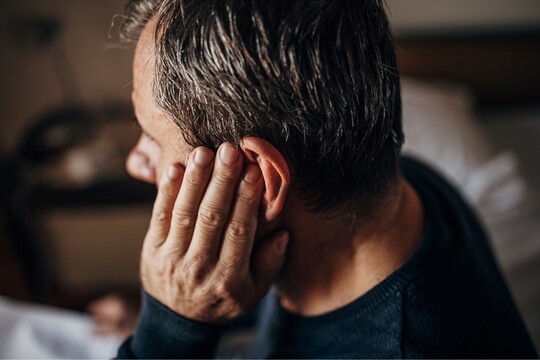 Ohrenschmerzen und ihre Ursachen – Entlastung für Hals und Stimme mit GeloRevoice® Mann hält sich das Ohr, möglicherweise aufgrund von Schmerzen oder einem Ohrproblem, in einem intimen, häuslichen Umfeld.