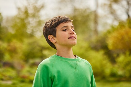 Junge im grünen Pullover steht draußen in einem Garten und atmet tief durch als Zeichen der Erholung nach Reizhusten.