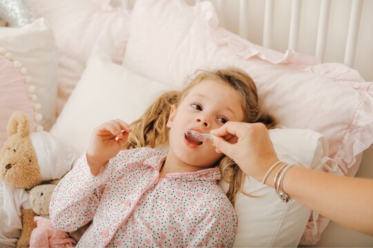 Krankes Mädchen im Bett, das von einer erwachsenen Person mit einem Löffel Medizin versorgt wird. Sie trägt Pyjama und hat Kuscheltiere neben sich.