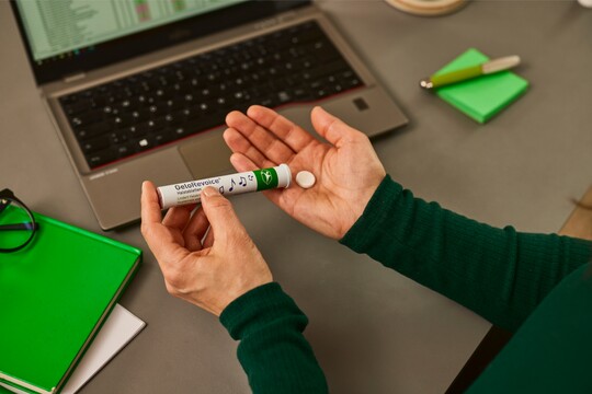 Eine Person hält eine GeloRevoice® Halstablette und die dazugehörige Verpackung in der Hand. Im Hintergrund befinden sich ein Laptop und grüne Notizbücher.