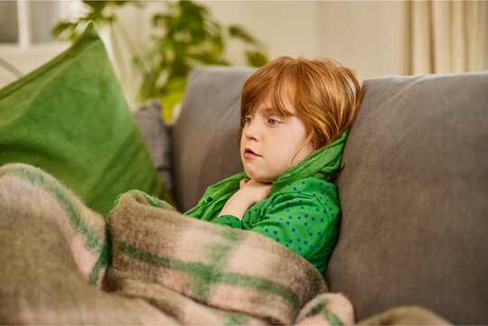 Ein junges Mädchen mit roten Haaren sitzt auf einem Sofa, in eine Decke gehüllt und hält sich den Hals, was auf Halsschmerzen oder Reizhusten hinweist.