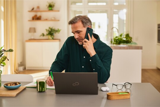 Ein Mann mittleren Alters sitzt in einem modernen, hellen Raum und telefoniert, während er sich Notizen macht. Vor ihm stehen ein Laptop und eine Brille. Die Szene vermittelt, dass er beruflich viel spricht und seine Stimme beansprucht, was auf die Notwendigkeit von GeloRevoice® zur Unterstützung bei Reizhusten und Heiserkeit hinweist.