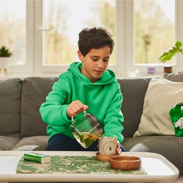 Junge in grünem Hoodie gießt Kräutertee aus einer Glaskanne in eine Tasse auf einem Wohnzimmertisch. Neben ihm liegt eine Packung GeloRevoice® Halstabletten, die für Unterstützung bei Halskratzen und Heiserkeit sorgt.