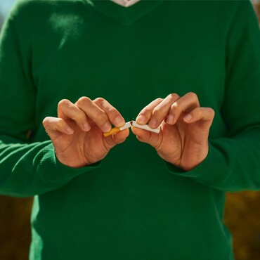 Raucherentwöhnung – Schutz vor Halsschmerzen und Mandelentzündungen. Eine Person in einem grünen Pullover bricht eine Zigarette in zwei Hälften. Die Szene symbolisiert das Aufhören mit dem Rauchen, um die Gesundheit des Halses zu fördern und Mandelentzündungen sowie Halsschmerzen zu vermeiden.