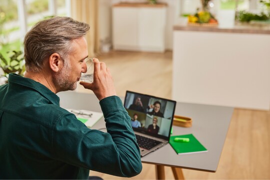 Mann trinkt Wasser während einer Videokonferenz für eine ausreichende Flüssigkeitszufuhr bei Rachenentzündung. Ein Mann trinkt ein Glas Wasser, während er an einer Videokonferenz auf seinem Laptop teilnimmt. Er sitzt in einem hellen Raum mit Pflanzen und Notizbuch vor sich. Die Szene betont die Bedeutung der Flüssigkeitszufuhr zur Linderung von Halsschmerzen oder einer Rachenentzündung.