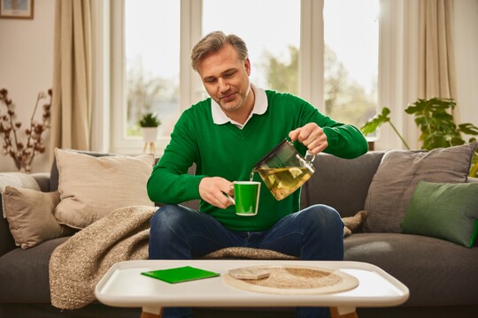 Ein Mann mittleren Alters sitzt auf einem Sofa und gießt sich eine Tasse Kräutertee aus einer Teekanne ein. Er trägt einen grünen Pullover, und im Hintergrund sind Kissen und Pflanzen zu sehen. Die Szene vermittelt die Anwendung von Hausmitteln wie Kräutertee zur Linderung von Reizhusten.