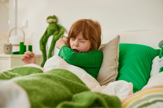 Mädchen mit roten Haaren und grünem Pullover liegt im Bett und hustet, umgeben von grünen Kissen und einem Plüschtier im Hintergrund.