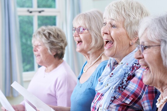 Eine Gruppe älterer Frauen singt gemeinsam in einem hellen Raum mit Fenstern. Sie halten Notenblätter in den Händen und lächeln während des Singens. Das Bild symbolisiert die Bedeutung von Stimmtraining im Alter zur Vorbeugung von Presbyphonie, die mit einer brüchigen und kraftlosen Stimme einhergeht.