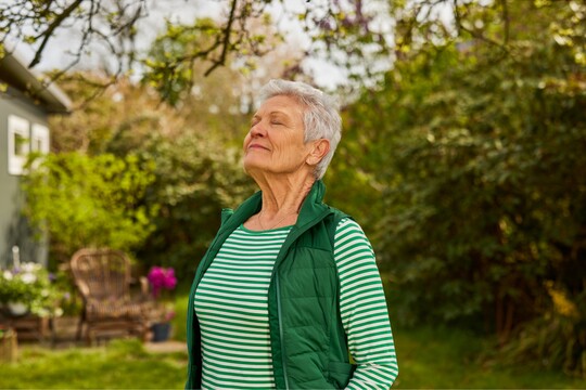 Ältere Frau in gestreiftem Oberteil und grüner Weste steht im Garten, schließt die Augen und atmet tief ein, umgeben von blühender Natur. Symbolisiert Entspannung und Atemübungen zur Stimmgesundheit.