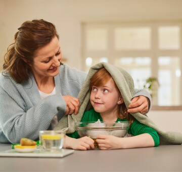 Eine Mutter hilft ihrem Kind beim Inhalieren über einer Schüssel mit heißem Wasser. Das Kind trägt ein grünes Oberteil und hat ein Handtuch über den Kopf gelegt. Auf dem Tisch steht eine Tasse heißes Wasser mit Zitronenscheiben. Die Szene zeigt ein traditionelles Hausmittel gegen Heiserkeit und Erkältungssymptome.