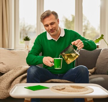 Ein Mann mittleren Alters sitzt auf einem Sofa und gießt sich eine Tasse Kräutertee aus einer Teekanne ein. Er trägt einen grünen Pullover, und im Hintergrund sind Kissen und Pflanzen zu sehen. Die Szene vermittelt die Anwendung von Hausmitteln wie Kräutertee zur Linderung von Reizhusten.
