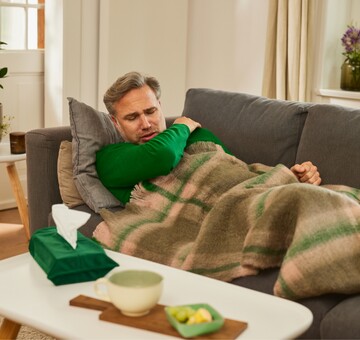 Ein Mann liegt krank auf einem Sofa, zugedeckt mit einer Decke, und hält sich den schmerzenden Hals. Auf dem Tisch vor ihm stehen eine Tasse Tee, Zitronenscheiben und eine Packung Taschentücher.