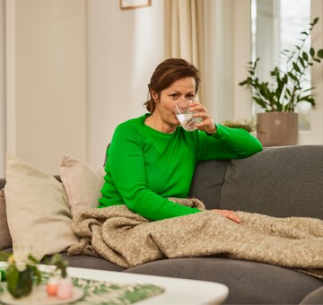 Frau in grünem Pullover sitzt auf einem Sofa und trinkt ein Glas Wasser. Sie ist in eine beige Decke gehüllt, und im Hintergrund stehen Pflanzen, was eine ruhige und entspannte Atmosphäre schafft. Die Szene deutet auf Stimmhygiene und die Wichtigkeit der Flüssigkeitszufuhr für eine gesunde Stimme hin.