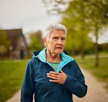 Ältere Frau im Freien, die besorgt ihre Hand auf die Brust legt, möglicherweise als Ausdruck stimmlicher Beschwerden im Alter. Die Szene thematisiert die Herausforderungen durch Presbyphonie, bei der die Stimme brüchig und weniger kräftig wird, oft begleitet von einem typischen Zittern des Kehlkopfes.