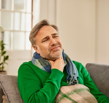 Mann mittleren Alters in grünem Pullover sitzt auf einem Sofa, hält sich den schmerzenden Hals und wirkt gequält. Er trägt einen Schal und scheint unter Heiserkeit oder Stimmverlust zu leiden.