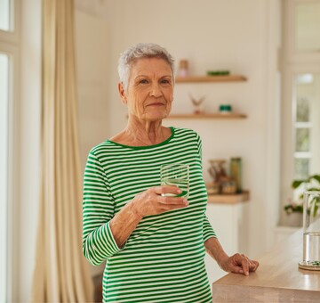 Ältere Frau in gestreiftem grünen Oberteil hält ein Glas Wasser in der Küche und blickt ruhig in die Kamera.