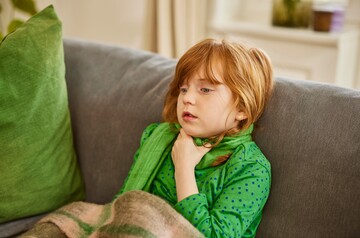 Ein kleines Mädchen mit roten Haaren sitzt auf einem Sofa und hält sich den Hals, während es einen Schal trägt. Sie wirkt erschöpft und hat offensichtlich Halsschmerzen. Eine Decke liegt über ihren Beinen, was auf Ruhe und Erholung hinweist.