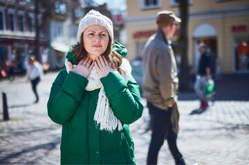 Eine Frau in einer grünen Winterjacke und mit Mütze hält sich auf einem belebten Platz in der Stadt die Hände an den schmerzenden Hals. Sie sieht besorgt aus, was auf Halsschmerzen oder eine Rachenentzündung hinweist.