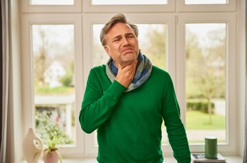 Ein Mann in einem grünen Pullover fasst sich mit schmerzverzerrtem Gesicht an den Hals, als Zeichen von Halsschmerzen, während er in einem hellen Raum mit großen Fenstern steht.
