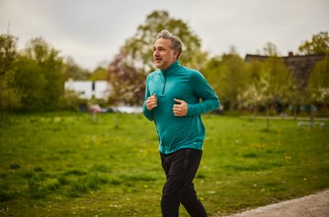 Ein Mann mittleren Alters joggt im Freien auf einem Pfad durch eine grüne Parklandschaft. Er trägt ein türkisfarbenes Langarmshirt und wirkt entspannt. Die Szene zeigt die Verbindung von sportlicher Aktivität und der Vorsorge bzw. Linderung von Halsschmerzen durch Bewegung.