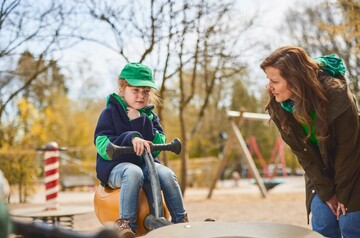 Kind sitzt auf einem Spielgerät im Park und hält sich den Hals, während eine Frau sich zu ihm herunterbeugt und besorgt schaut.