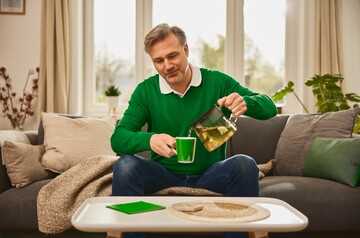Ein Mann mittleren Alters sitzt auf einem Sofa und gießt sich eine Tasse Kräutertee aus einer Teekanne ein. Er trägt einen grünen Pullover, und im Hintergrund sind Kissen und Pflanzen zu sehen. Die Szene vermittelt die Anwendung von Hausmitteln wie Kräutertee zur Linderung von Reizhusten.