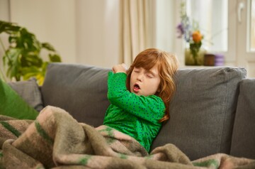 Ein rothaariges Mädchen hustet in ihren Ellenbogen, während es auf einem Sofa sitzt, in eine Decke gehüllt.