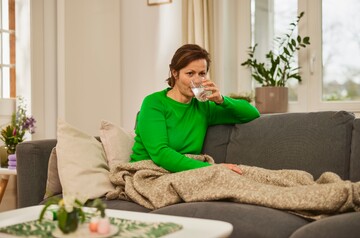 Frau in grünem Pullover sitzt auf einem Sofa und trinkt ein Glas Wasser. Sie ist in eine beige Decke gehüllt, und im Hintergrund stehen Pflanzen, was eine ruhige und entspannte Atmosphäre schafft. Die Szene deutet auf Stimmhygiene und die Wichtigkeit der Flüssigkeitszufuhr für eine gesunde Stimme hin.